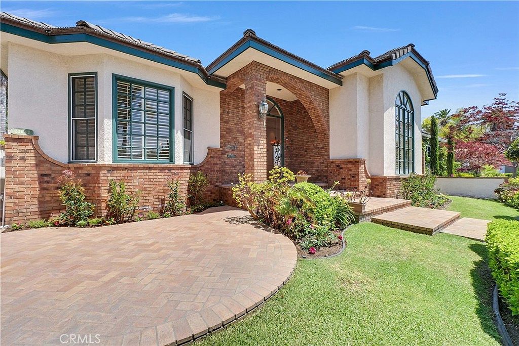 A brick-paver driveway curving toward the home, lined with planted beds, leading to a brick-arched portico and tall arched windows trimmed in teal