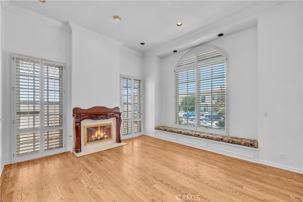 The main house living room, a white-walled high-ceilinged volume with refinished hardwood floors, a carved wood-mantel fireplace lit with a gas flame, plantation shutters on tall doors flanking the mantel, and a tall arched plantation-shuttered window over a built-in upholstered window seat