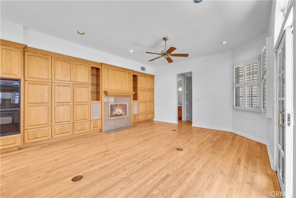 The family room of the main house, a wall of oak built-in cabinetry framing a tiled fireplace lit with a gas flame, a ceiling fan overhead, plantation-shuttered French doors to the side, all on refinished oak hardwood