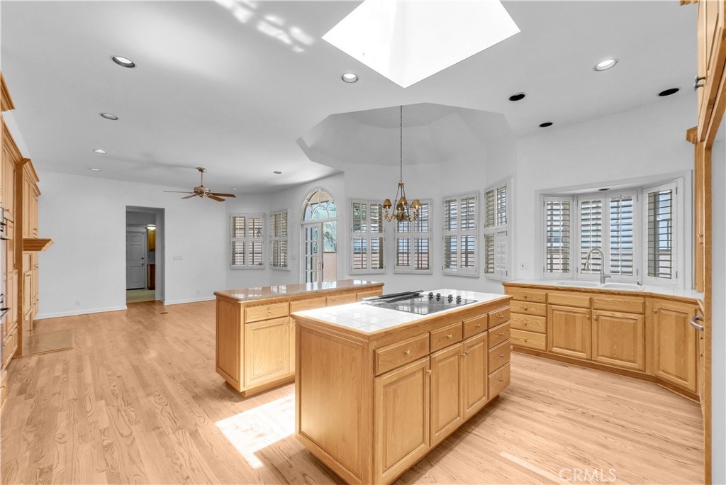 The kitchen of the main house, a vaulted-ceiling room with a pyramidal skylight at the peak, two islands in oak cabinetry with tile counters, plantation-shuttered bay windows along the back wall, and a chandelier over the eat-in dining area beyond