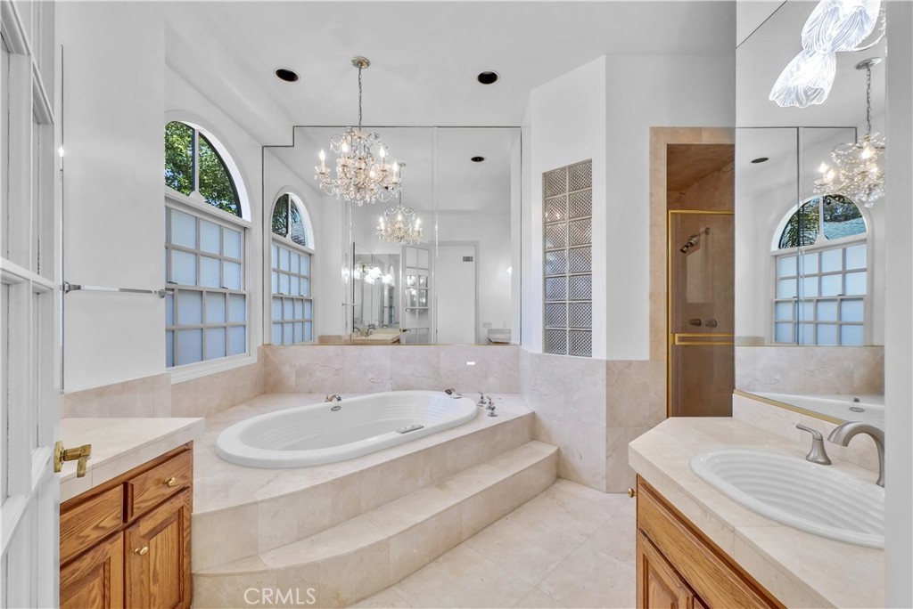 The primary bath, a stone-tiled room with an oval soaking tub on a stepped platform under a chandelier, framed by a wall of mirror, two vanities flanking the tub, glass-block windows, and a separate glass-walled shower beyond
