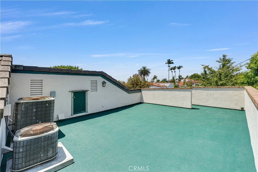 The rooftop deck above the upper guest unit, a wide flat teal-painted deck wrapping around a small mechanical hutch and a service door, low stucco walls along the perimeter, palms and Belmont Heights rooftops visible beyond, a wide flat blue Long Beach sky overhead